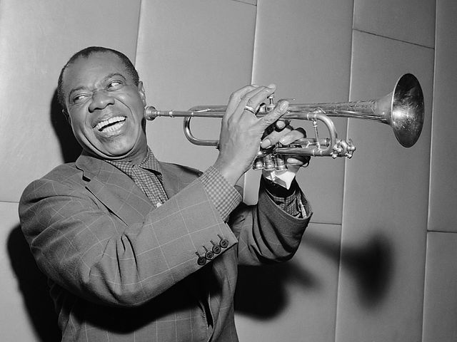 Louis Armstrong is shown in black-and-white smiling as he turns his head away from his trumpet. He holds his trumpet up to his face.