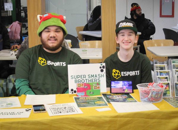 Two student members of WCC's E-sports Club sit at a table to advertise for new members. They are both smiling, wearing hats, and green E-sports t-shirts