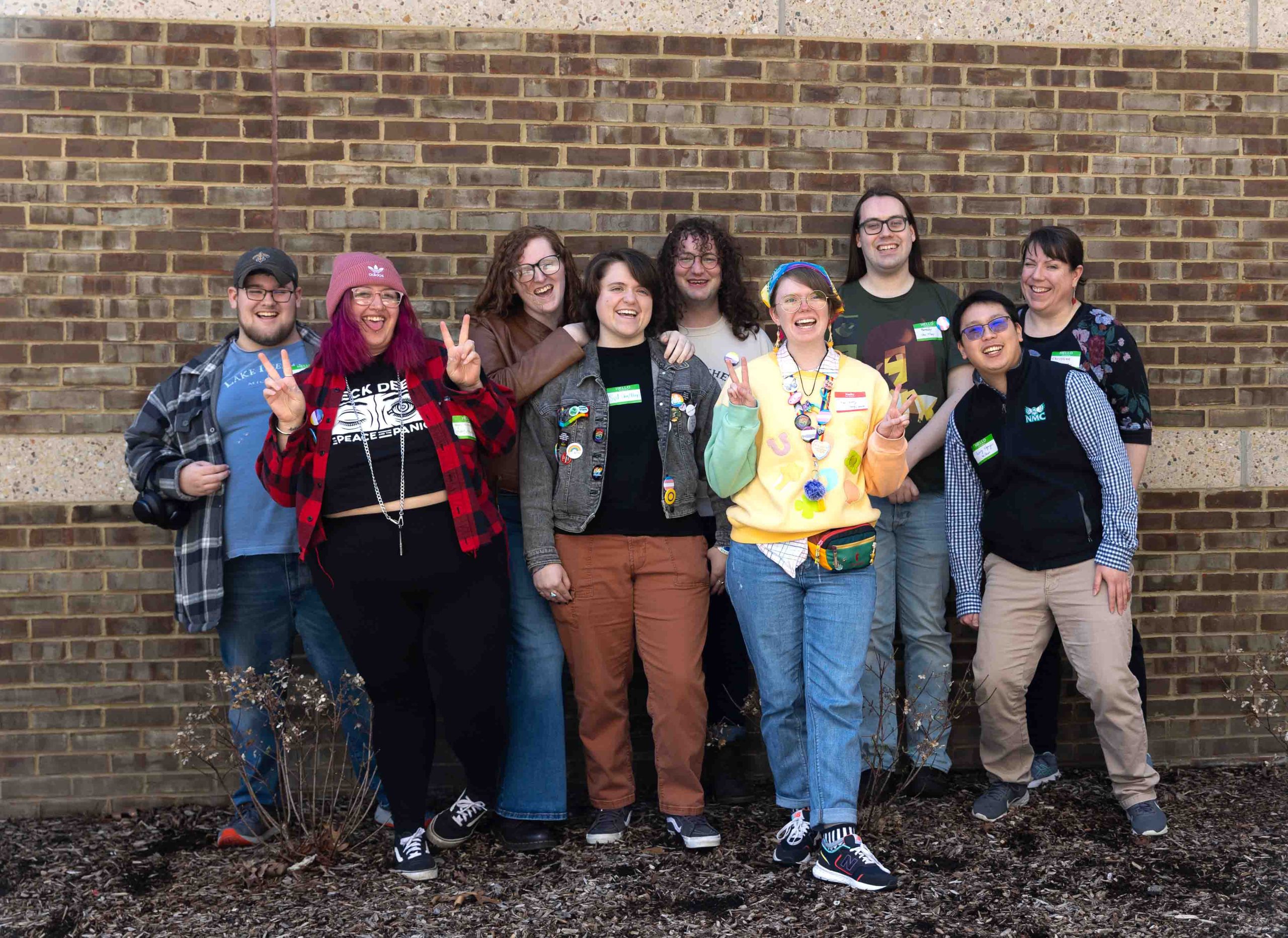 A group of nine students pose in front of a brick background while smiling and having a good time