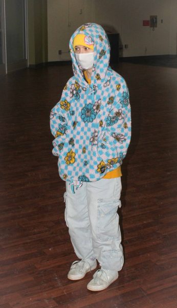 Ingrid Lamble wears the hood up on a blue, white, & yellow fuzzy hoodie with checks & flowers & light blue denim cargo pants.