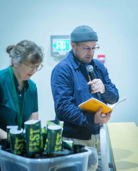 Matt Zacharias stands reading from a book and speaks into a microphone while announcing the Vid Fest winners