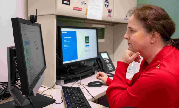 Barbara Hauswirth, wearing a bright red sweater, rests her chin on her hand as she sits at her desk and peers at her computer screen