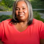 Headshot of Yana Mcguire, Staff Editor for the Washtenaw Voice, from the shoulders up, smiling and wearing a red shirt.