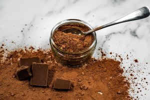 Cocoa powder from a jar spills over a marble countertop. A silver spoon rests inside the jar, and blocks of chocolate sit on the counter on the left.