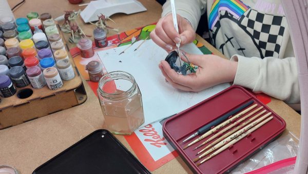 A close-up of someone painting a miniature figurine. Only their hands are visible. The table is covered with art supplies.
