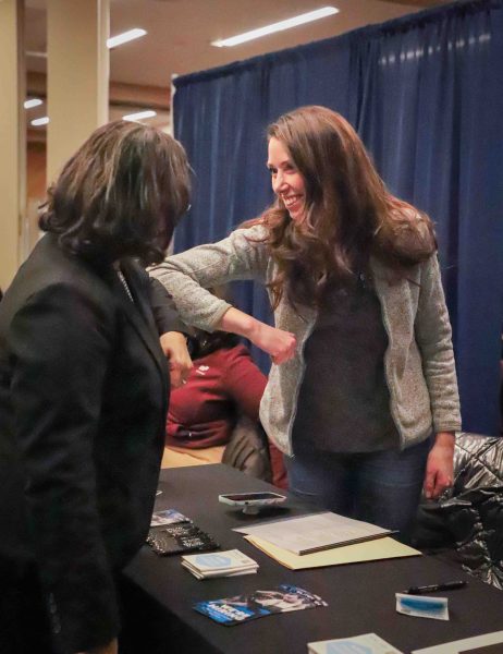 A participant and a representative are shown smiling and bumping elbows to greet each other.