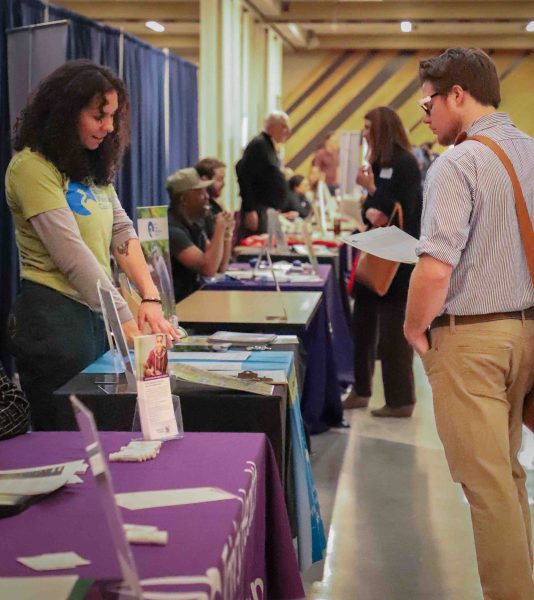 This photo of the career fair shows many tables lined up. They have various people sitting and standing at the tables.