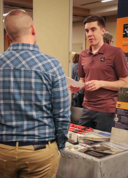 A student stands in front of a table, their back facing the camera. A representative stands behind the table.