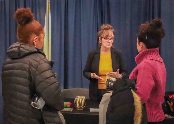 Two participants listen to a representative as she stands and speaks behind a table.