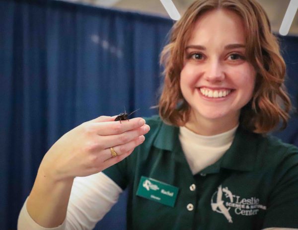 Rachel Lorenc, wearing a green T-shirt with a white long-sleeved shirt underneath, smiles while holding a cockroach.