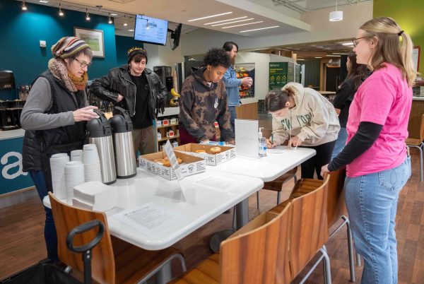 A table with coffee canisters, donuts, plates, cups and utensils is shown with students gathering around.