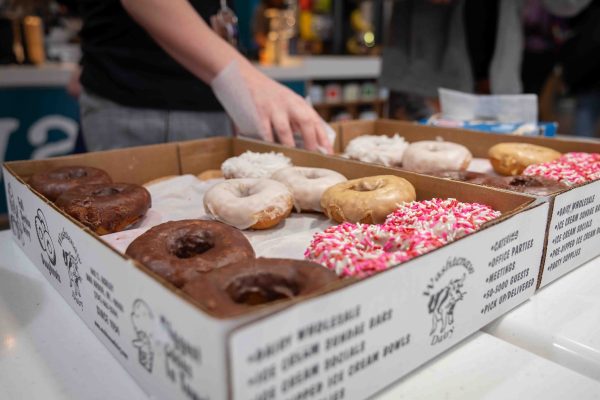 A close-up of a box of donuts is shown with someone grabbing one. Pink, white, glazed, and chocolate donuts are shown.