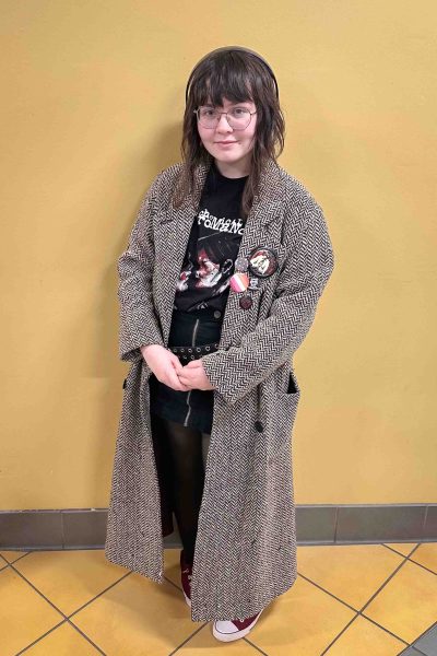 Photo of Eliza Atkins standing in front of a yellow wall, wearing a black-and-white coat over a My Chemical Romance T-shirt.
