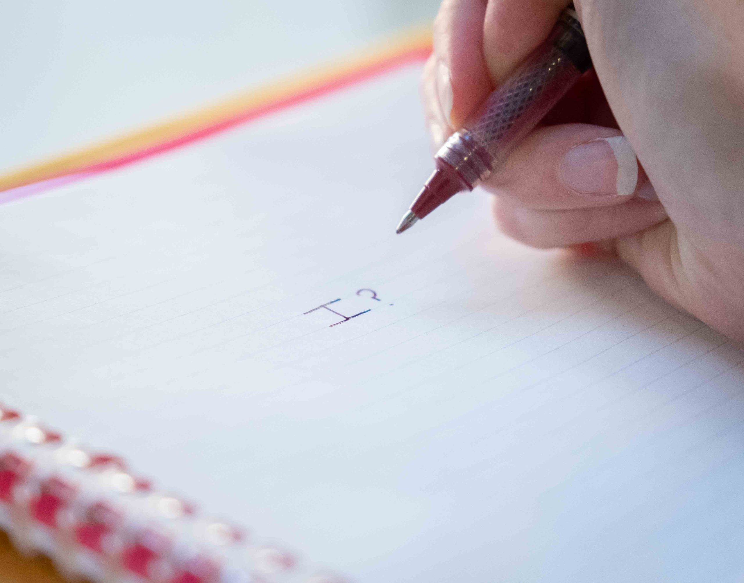 A close-up of someone writing "I?" on an otherwise blank page in a spiral notebook.