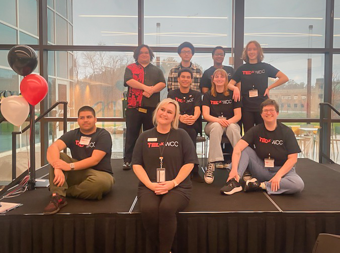 A group of students poses on a black platform stage for a group photo. They are all wearing TEDX T-shirts.