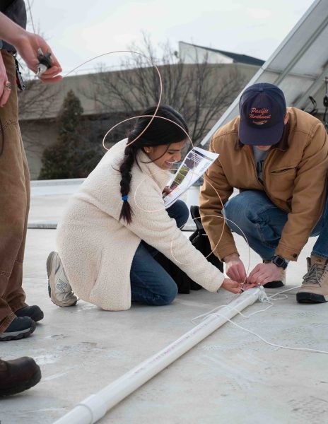 Two students are shown kneeling next to PVC pipe. Sruthilaya Rajavelu-Mohan smiles, copper wire stretched in front of her.