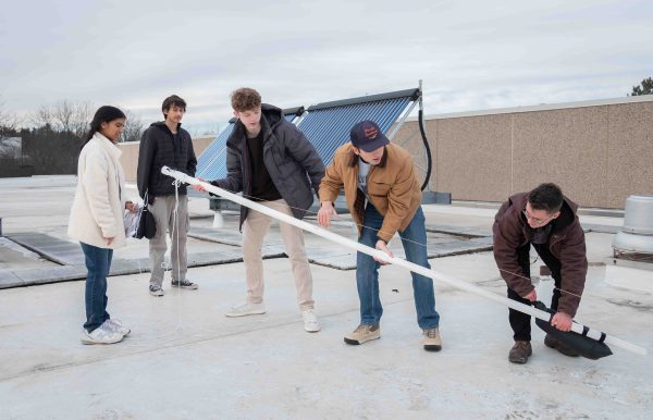 Three students hold a PVC pipe, leaning it upward slowly from the base. Two other students watch from the side.