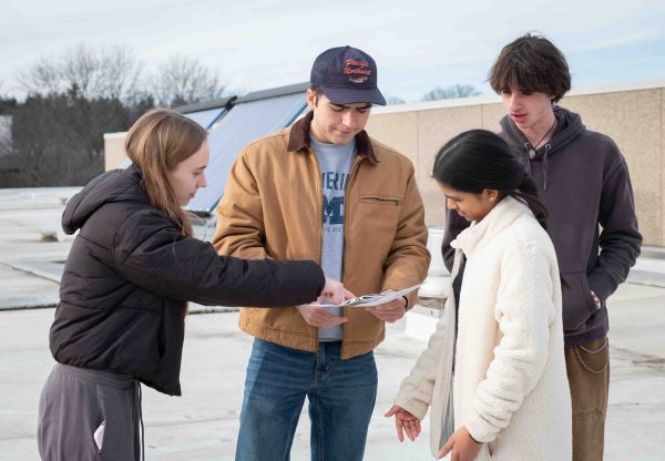 Four students study instructions from a booklet. The leftmost student is pointing to something in the instructions.