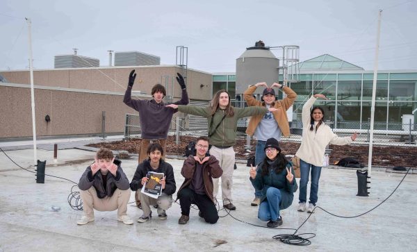8 students pose together in two rows. The front row kneels while the back row shapes the letters "WTMC" with their arms.