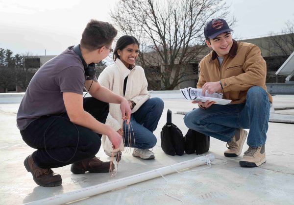 Three students crouch around a PVC pipe and smile while brainstorming. Owen Rian-Farley, left, holds a loose roll of wire.