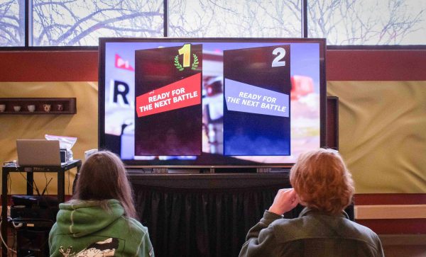 Jeyome Howell and Ethan McElmeel sit in front of a TV screen with backs to the camera. The screen has a video game starting.