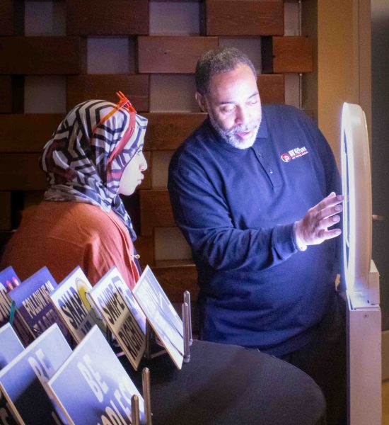 Jana Aboud sits in front of a large ring light while Mario Clark taps on the light and explains the photo-taking process.