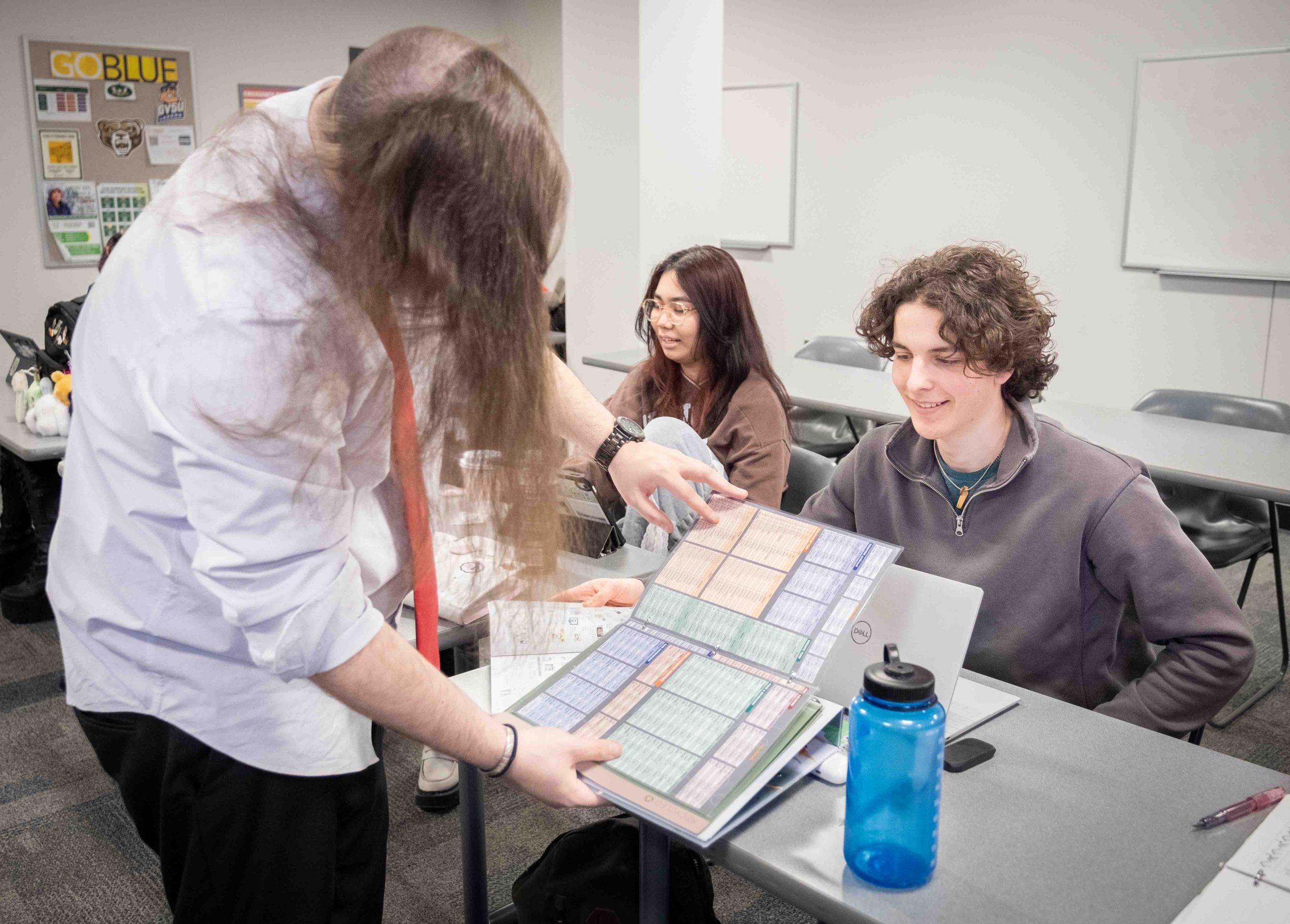 Two students sit at a table while another student stands in front, holding a book open toward the camera.