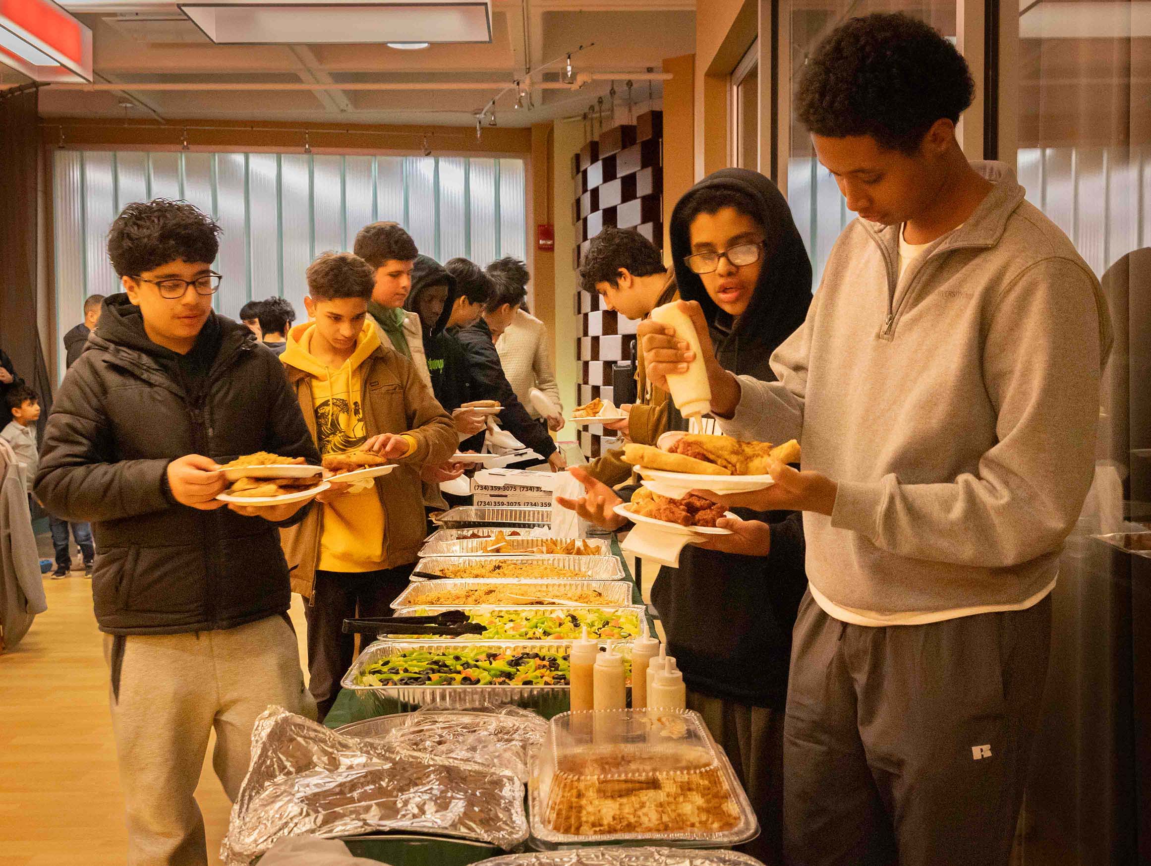 Male Muslim students fill their plates as they line up on both sides of a table filled with delicious food.