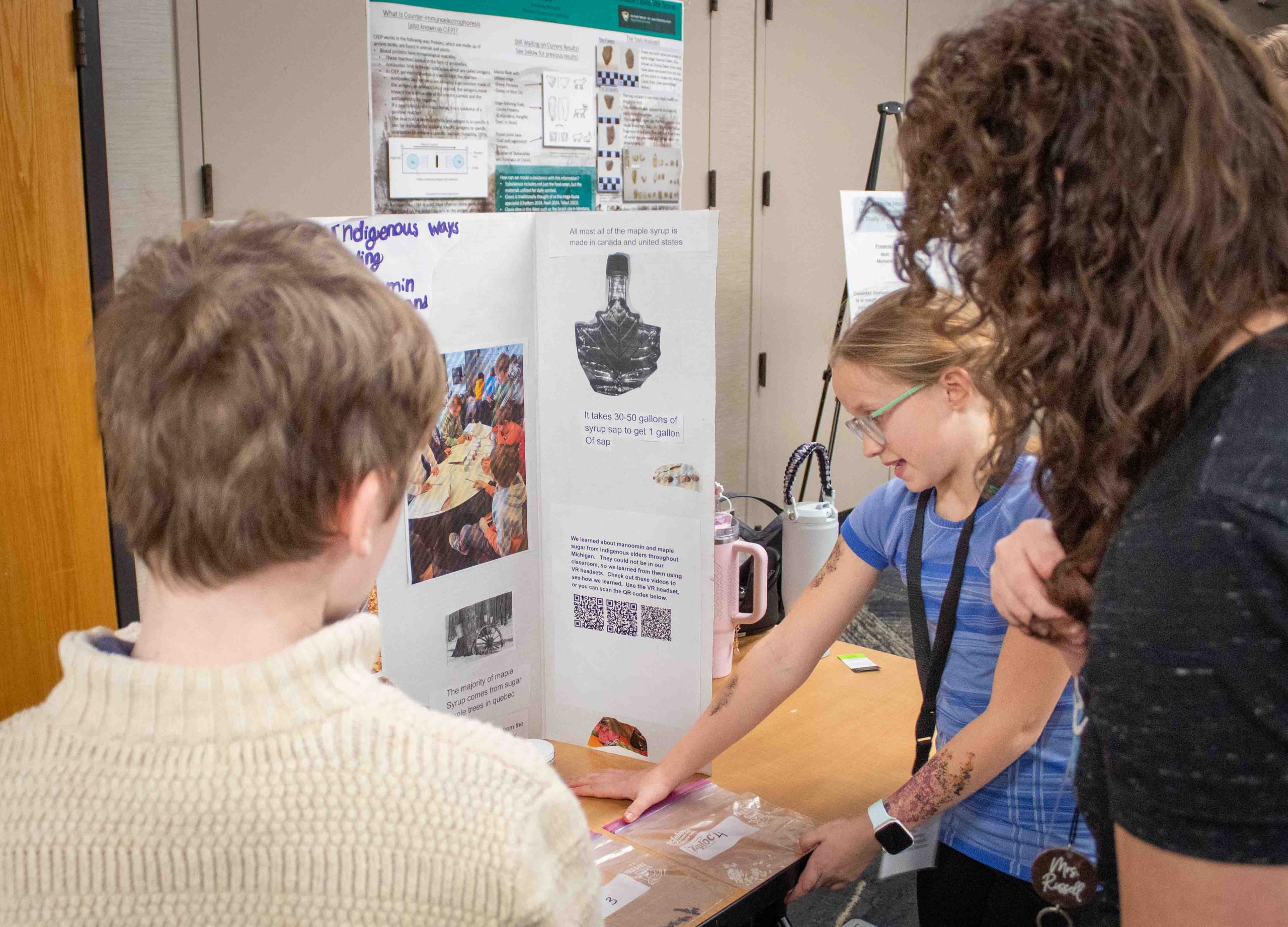 Two students present their poster to an attendee. One wears a blue shirt and the other wears beige.