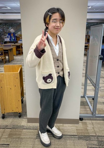 Sunny Park makes a peace sign while wearing a cream teddy bear cardigan over a brown vest and white collared shirt.