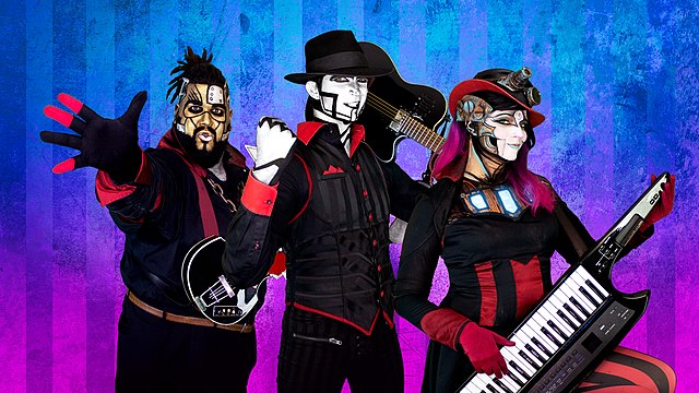 The members of Steam Powered Giraffe wear heavy makeup & hold instruments while standing in front of a blue & purple wall.