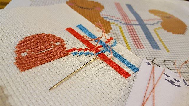 A golden needle rests on a colorful cross-stitched pattern.