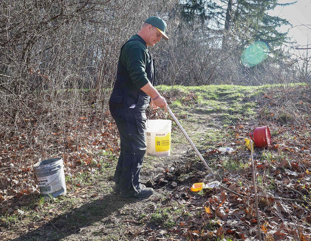 Ron Shulz, wearing a green shirt and overalls, uses a grabber to pick sticks off of the ground and put them into a bucket.