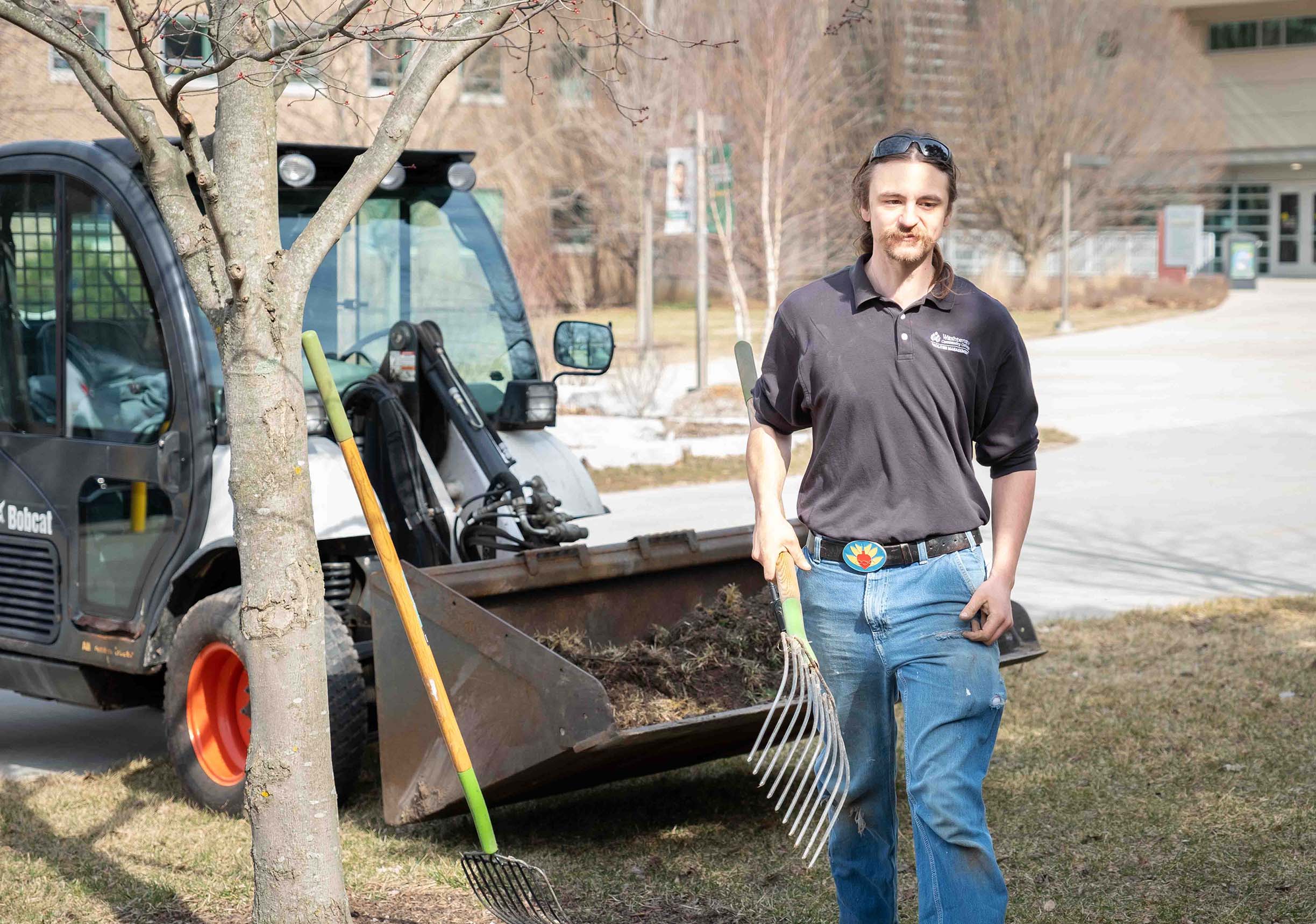 Caleb Horvath stands in front of a tree and Bobcat vehicle while holding a rake and wearing a black WCC T-shirt and jeans.