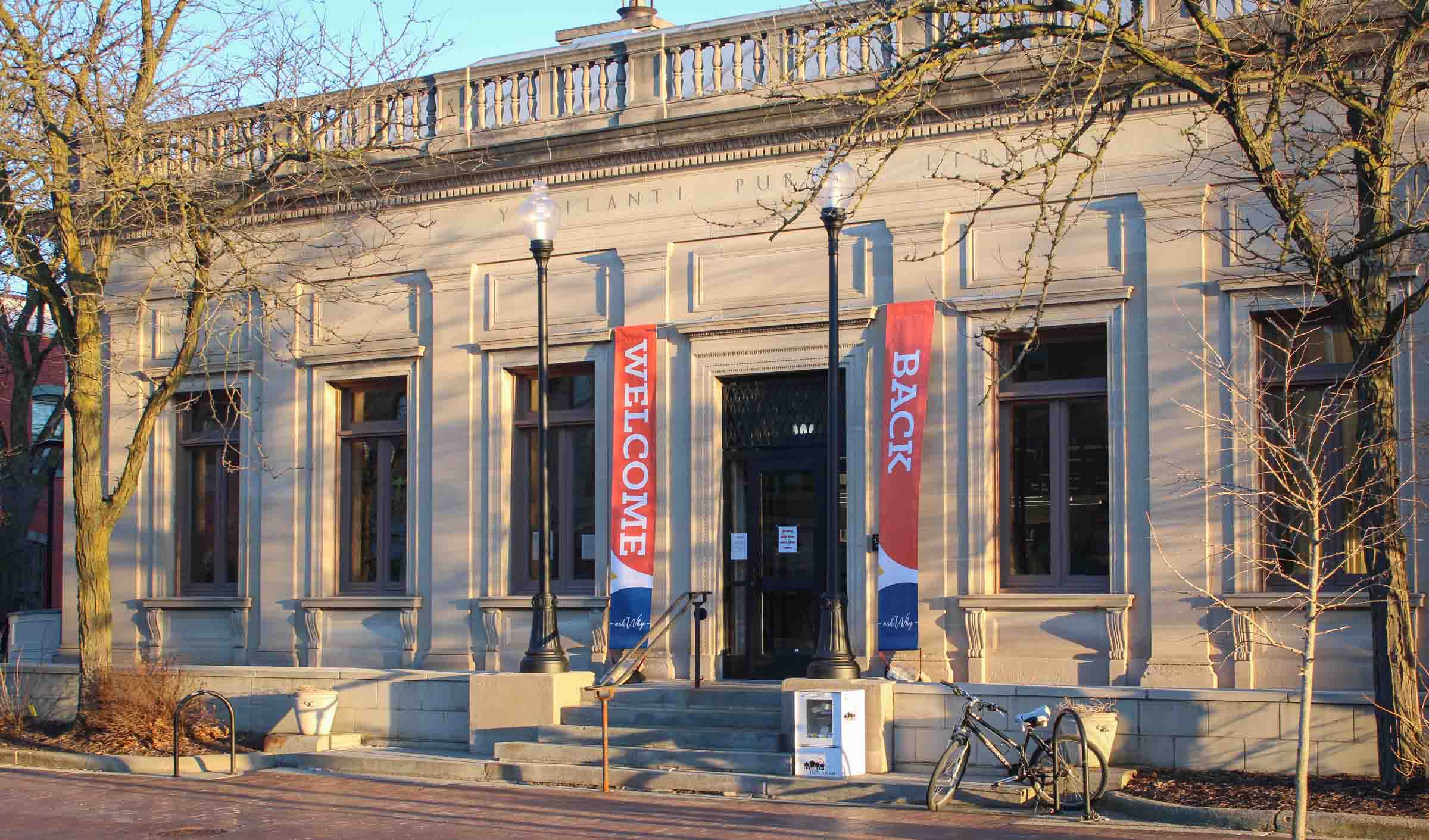 The Ypsilanti Community Library is a tan stone building with vintage architecture. "Welcome Back" banners hang by the door.