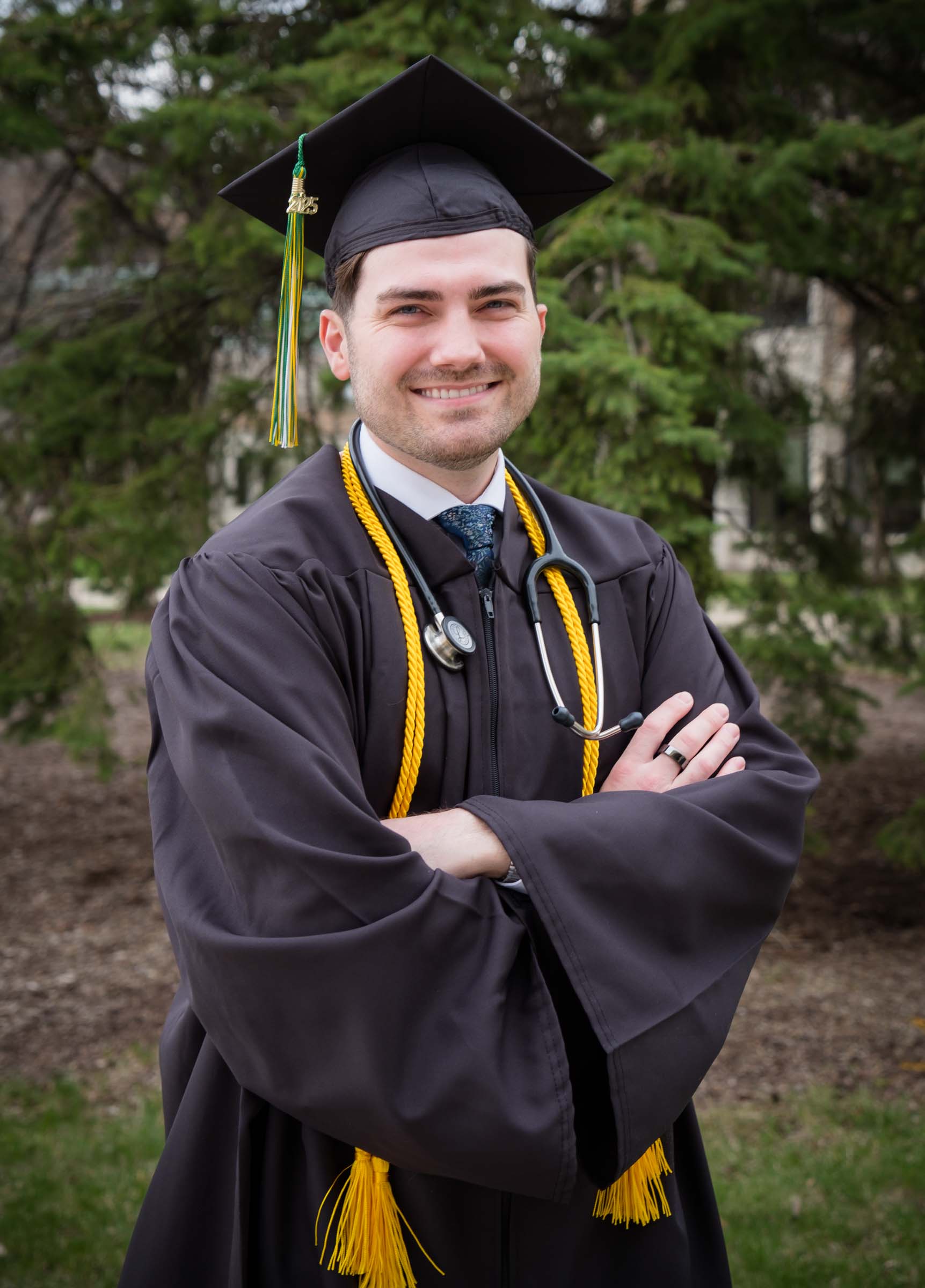 Bryan Wilson is shown from the waist up smiling and wearing graduation robes. A stethoscope is worn around his neck.