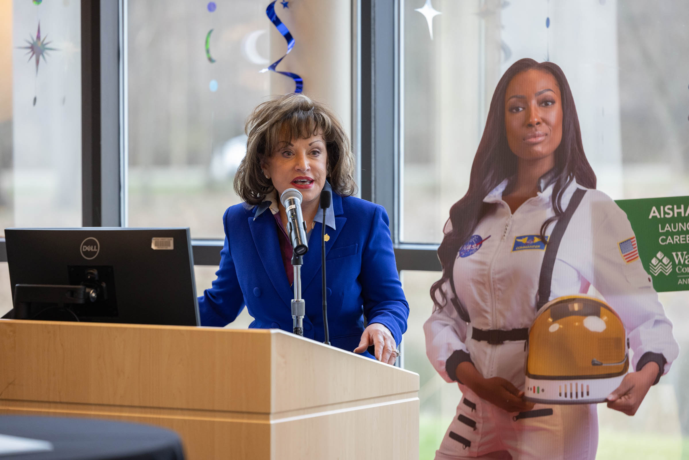 Rose Bellanca stands behind a wooden podium and speaks to the audience into a microphone with a cardboard cutout of Aisha Bowe standing behind her.