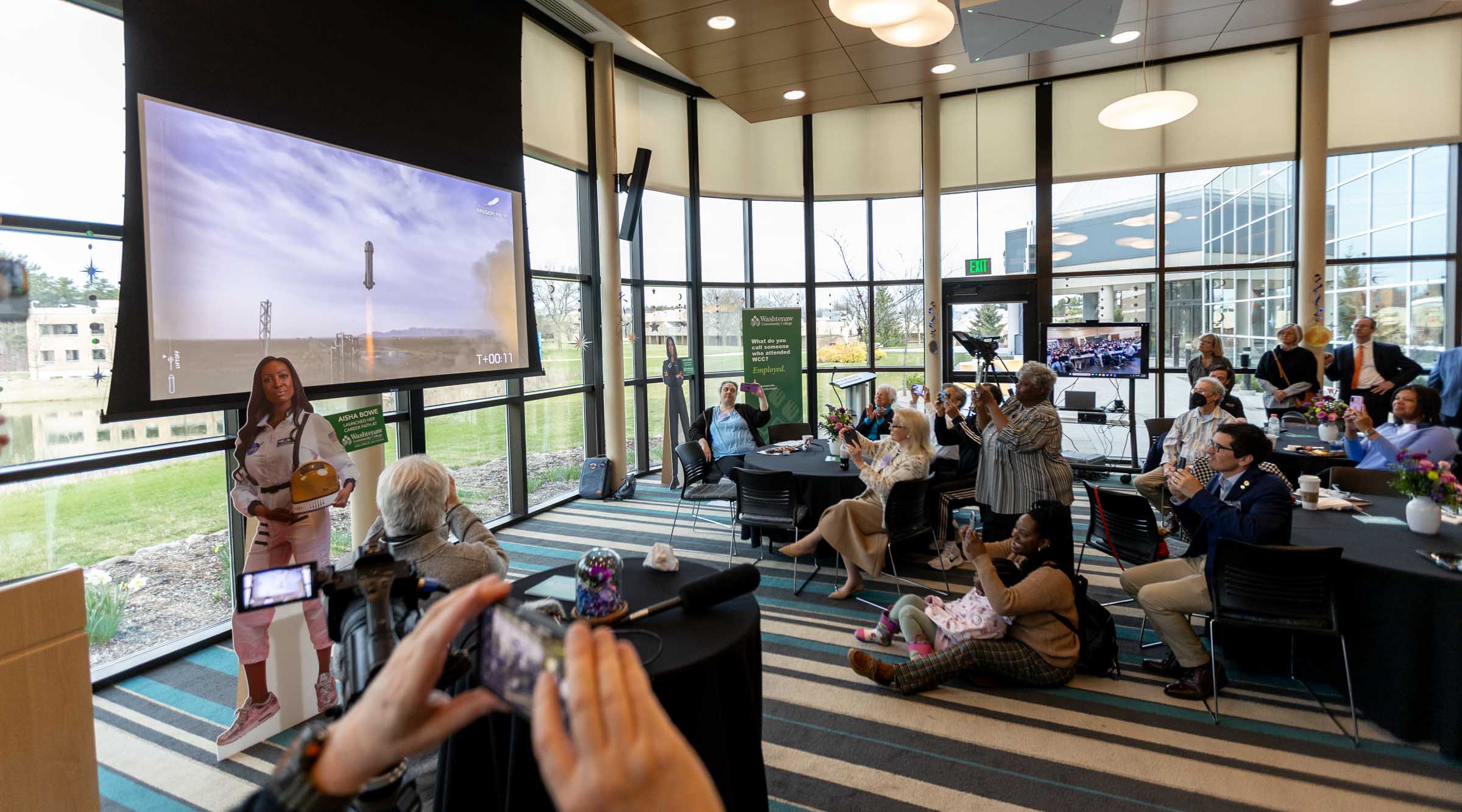 The rocket is shown taking liftoff on the big screen while attendees watch in excitement, many filming from their phones.