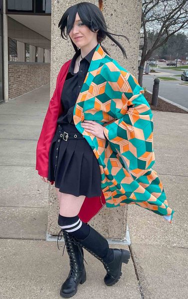 Samantha Peach wears a black wig, black shirt, and pleated skirt. A teal and orange haori jacket is layered over the outfit.