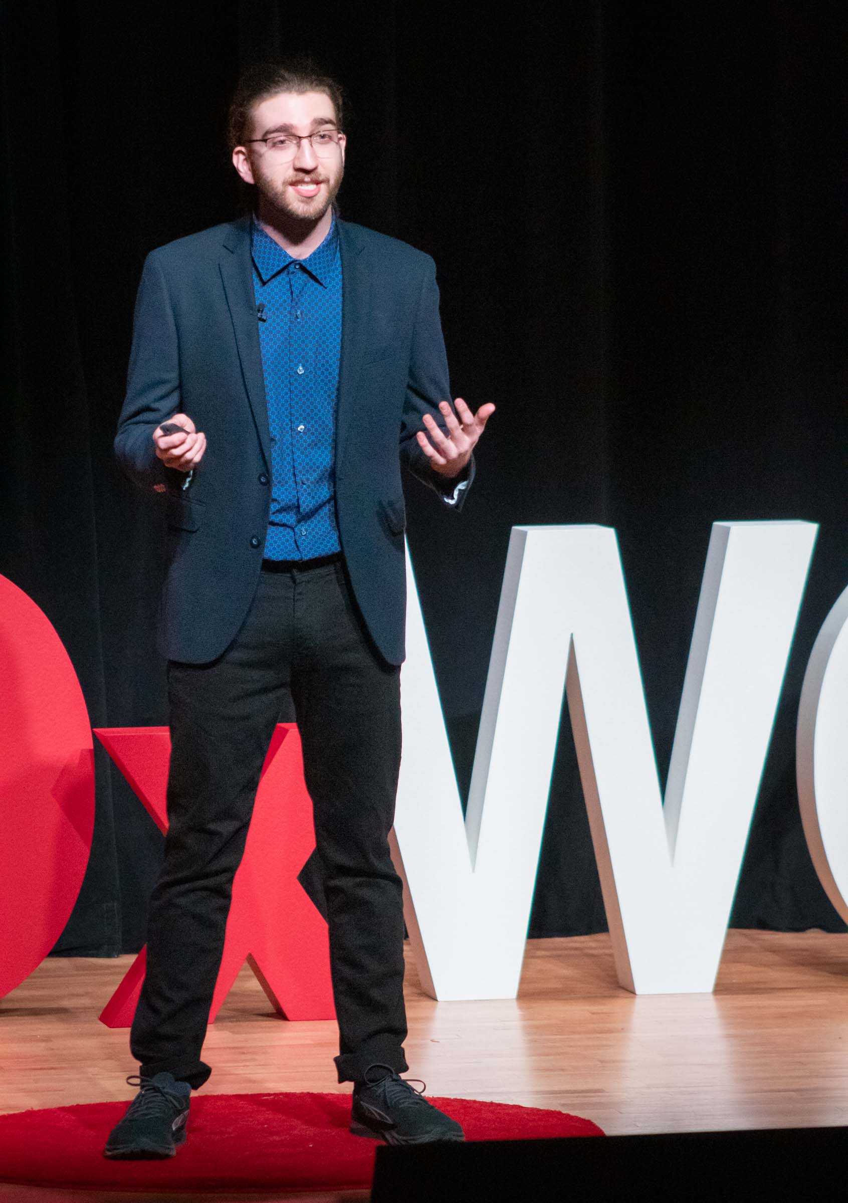 Dylan Orsolini speaks to the audience with his hands outstretched. He wears a dark suit with a blue dress shirt.