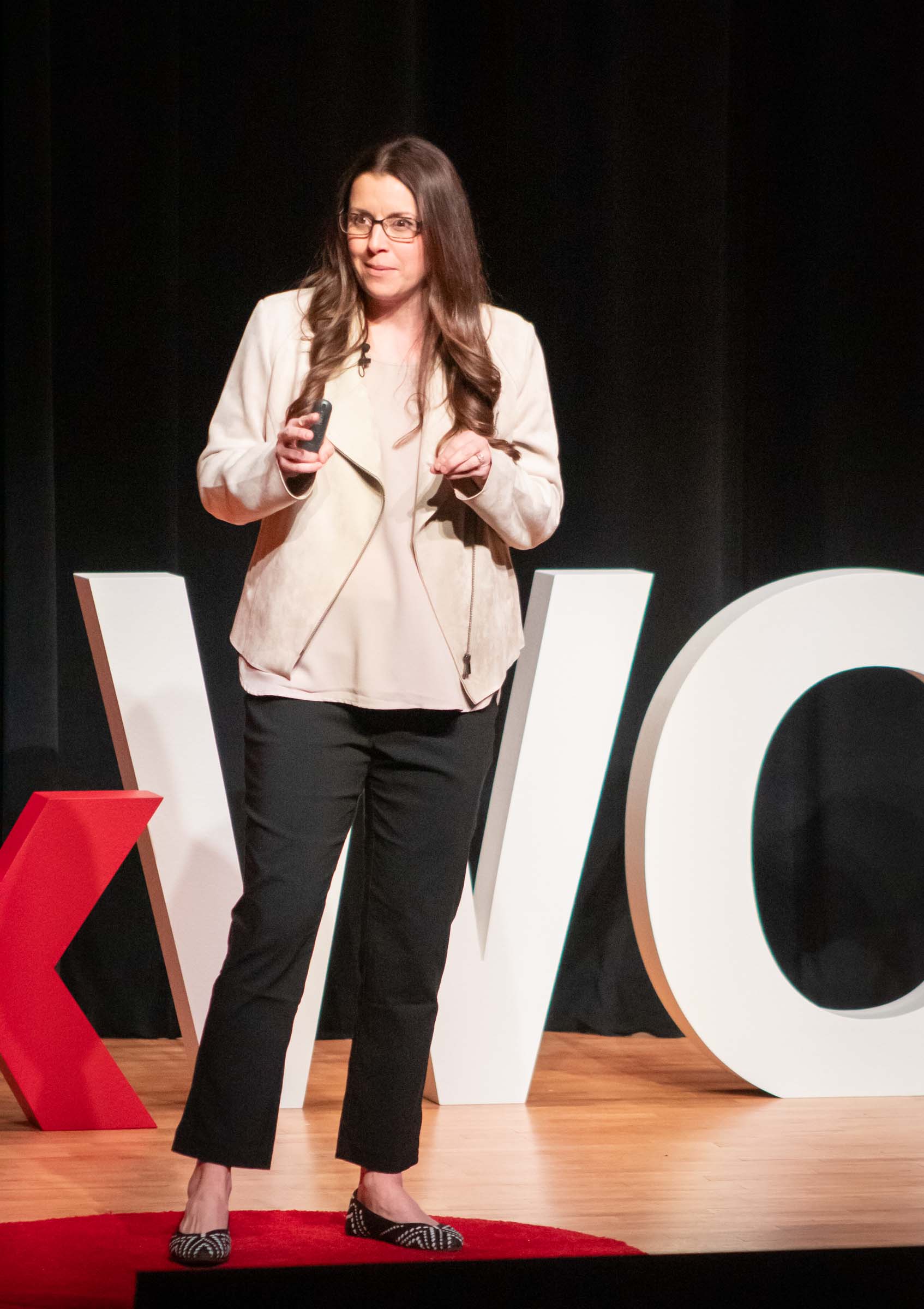 Jessica Hale stands on stage while speaking to the audience. She wears black slacks and a tan blazer.