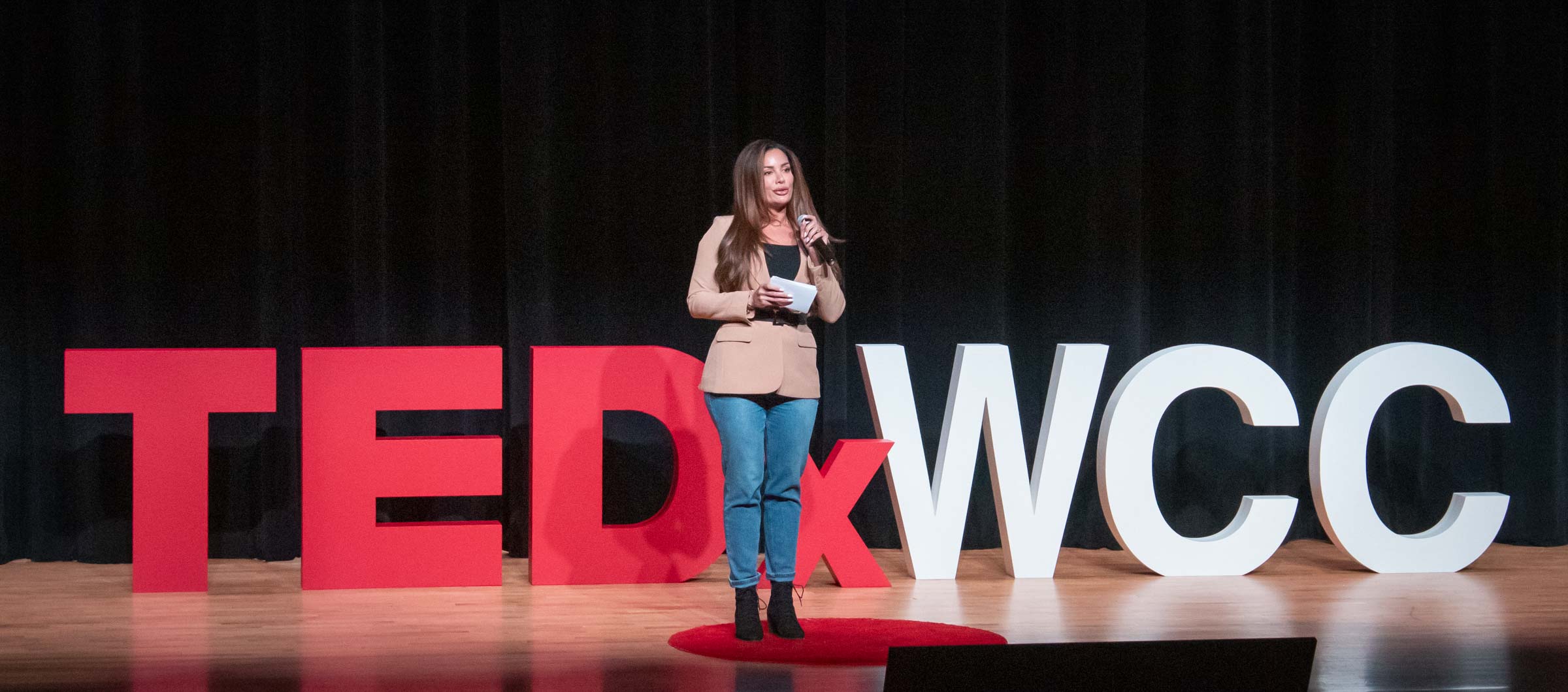 Rayan Salam stands onstage in front of the large lettered sign "TEDxWCC." She wears blue jeans and a beige jacket.
