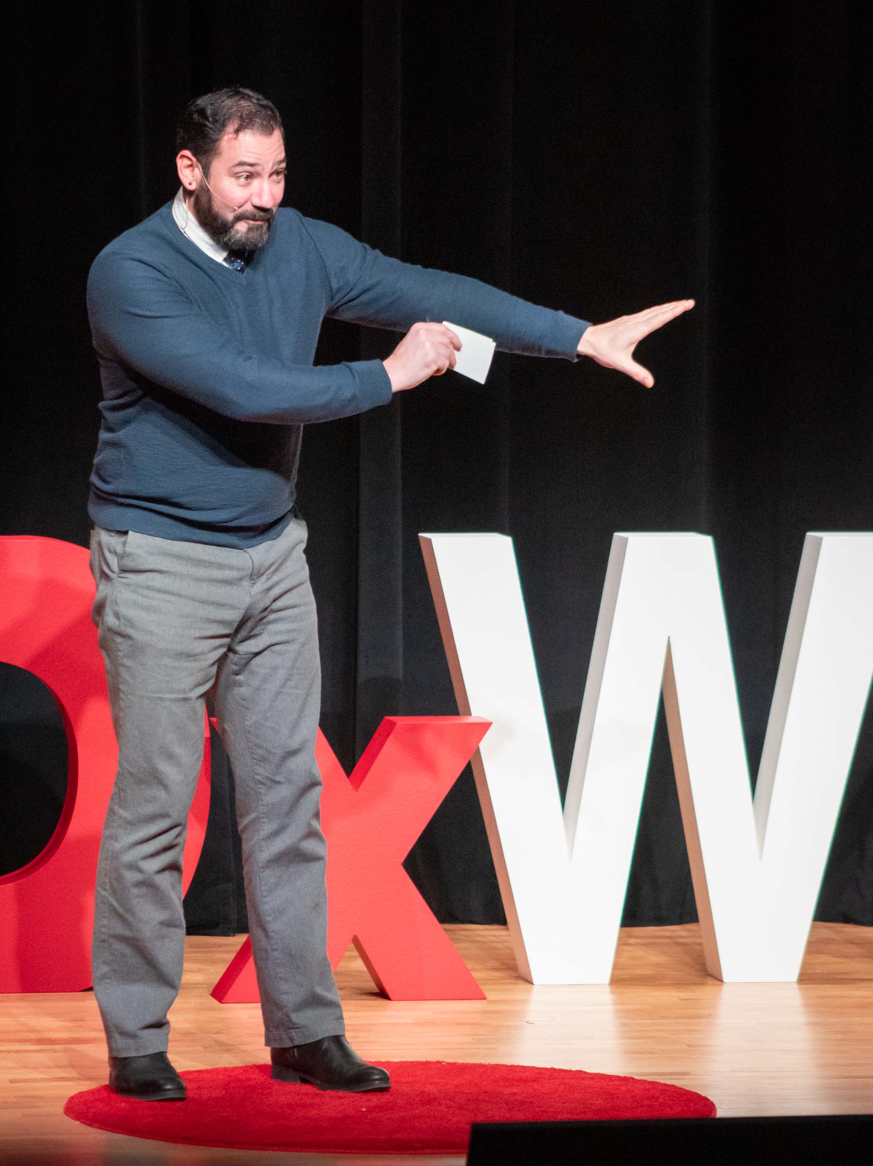 Jason Thompson speaks to the audience with his arms outstretched to the side. He wears a blue sweater with gray pants.