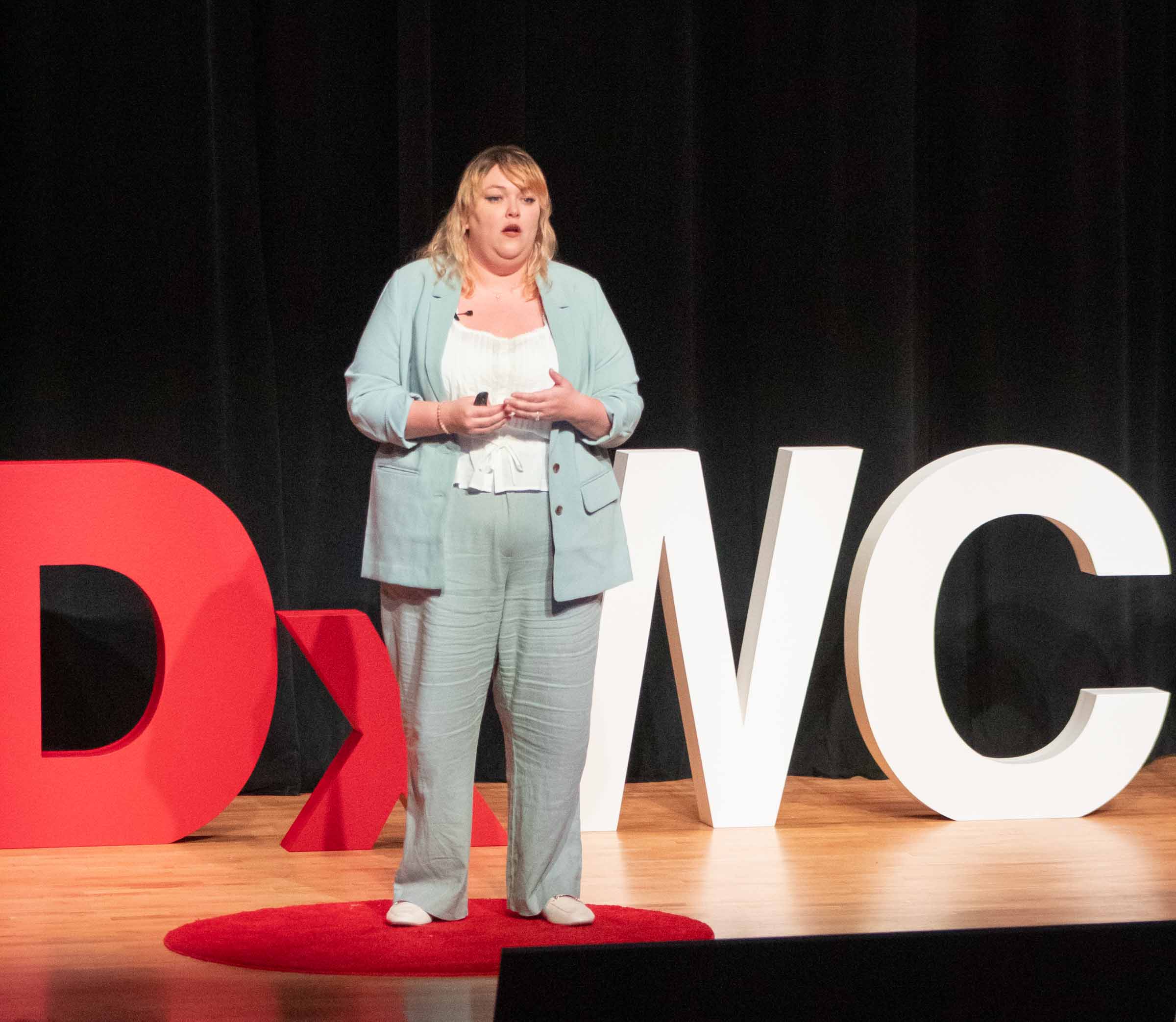Lukas Hendricks stands on stage and speaks to the audience, holding her hands at her waist. She wears a light blue suit.