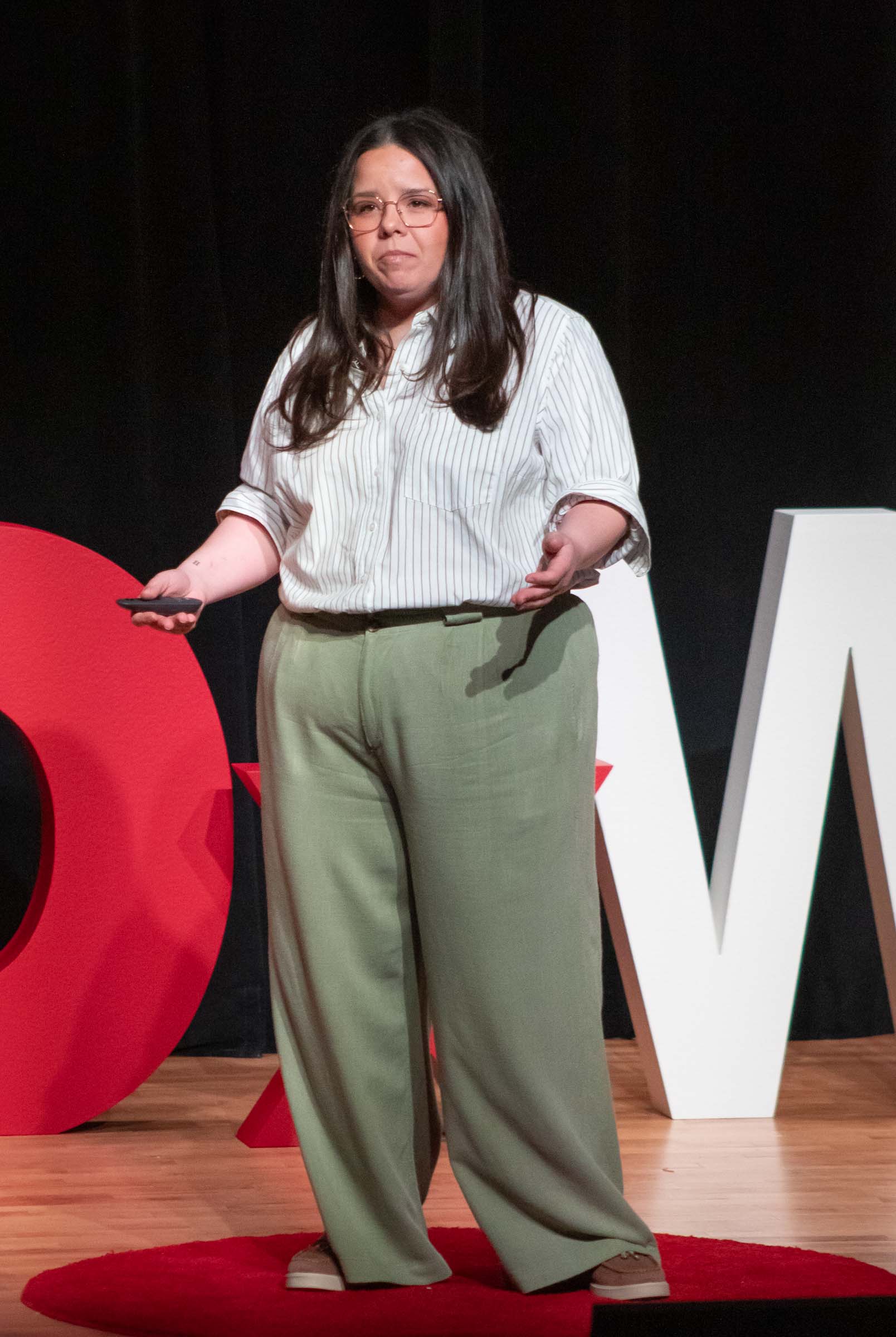 Kristina Moreno stands on the stage while speaking and gesturing to the audience. She wears a white shirt and green pants.