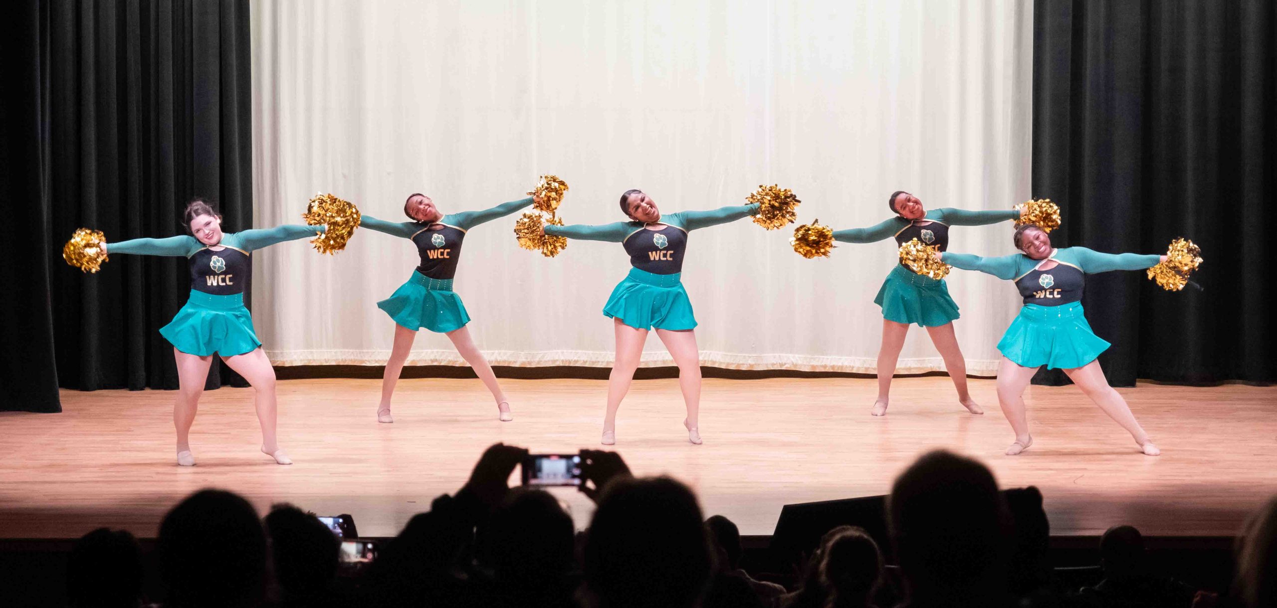 The WCC Dance Team performs on stage with black and green outfits and gold pom-poms.