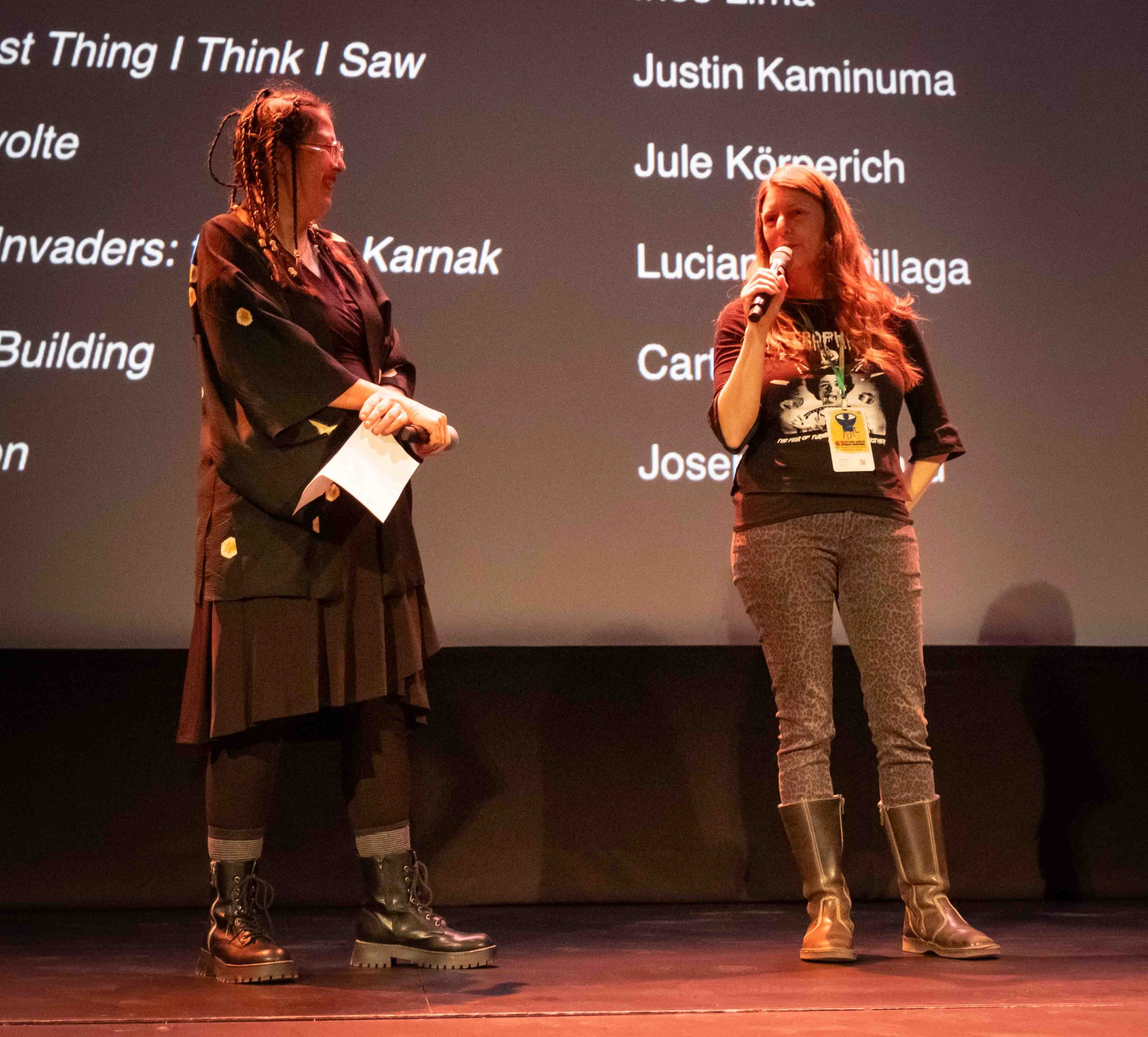 Leslie Raymond stands on stage with Jule Koperich and watches Jule speak into the microphone.