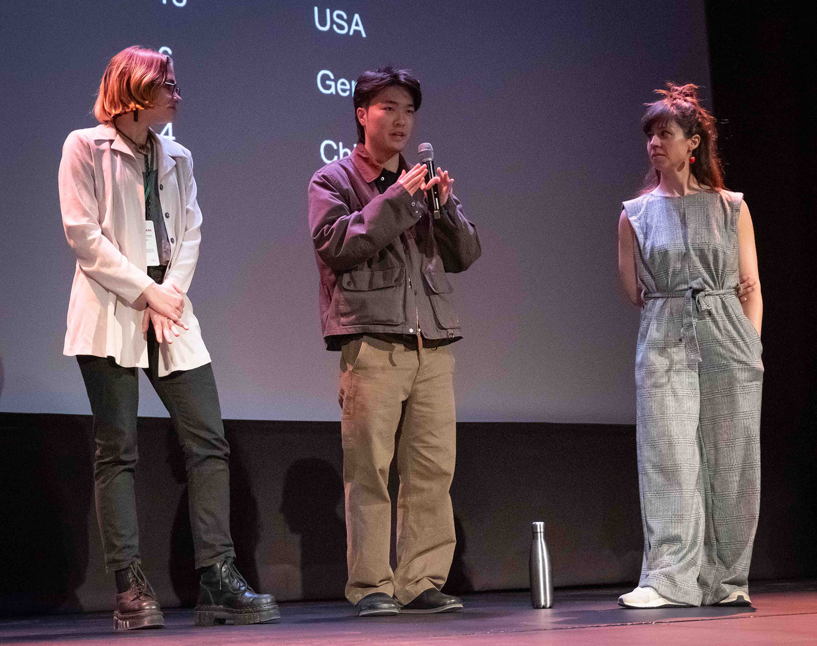 Three people stand on stage in a line. The outermost people watch Justin Kaminuma speak to the audience through a microphone.