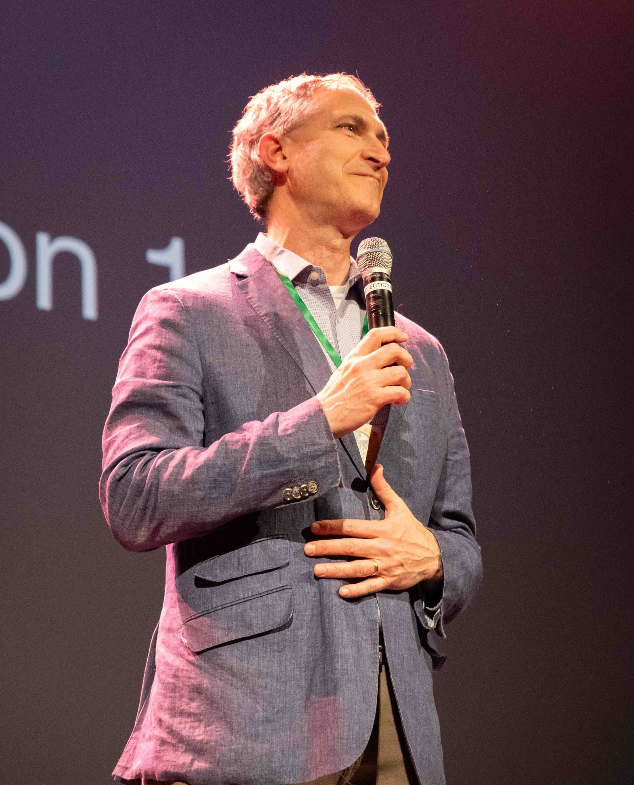 Christopher Taylor is pictured from the waist up wearing a grey suit. He grins and holds a microphone in his hand.