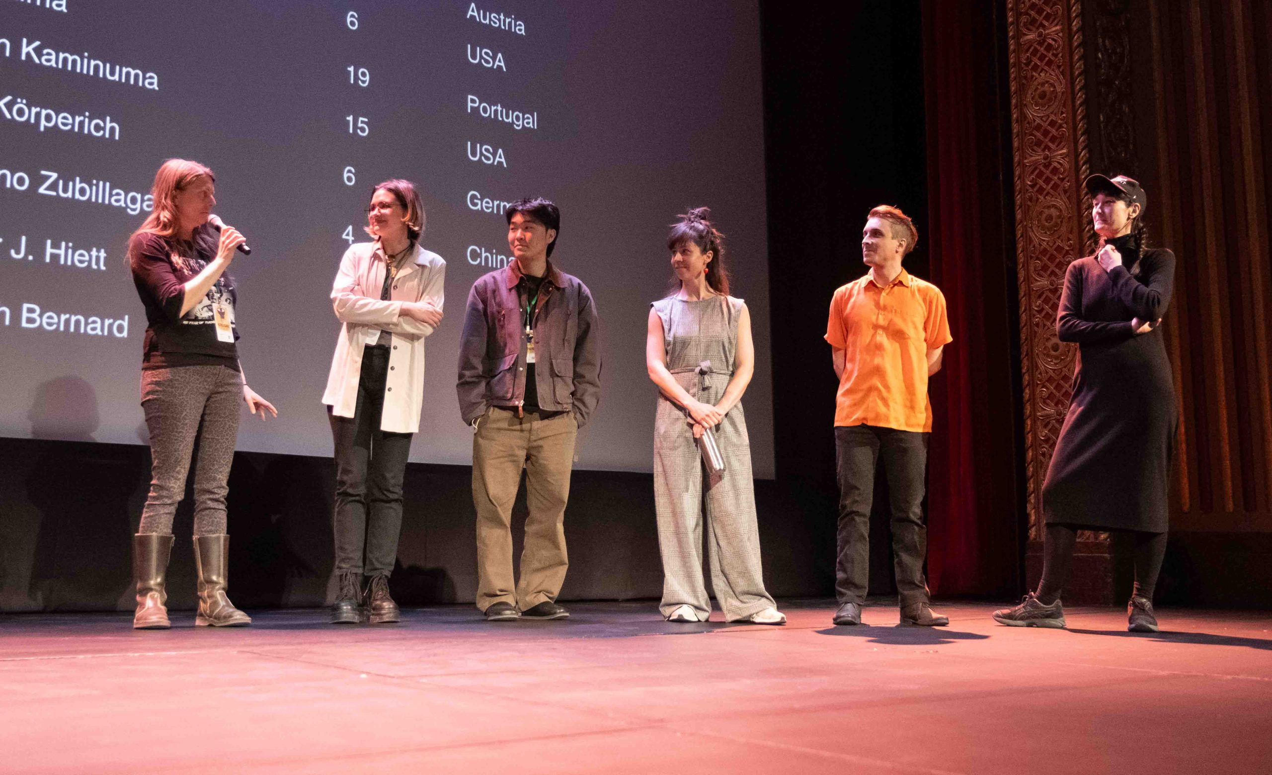 Six people stand on stage and watch Jule Korperich speak to the audience through a microphone.